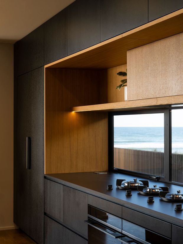 A refined contemporary kitchen with dark textured cabinetry, a black benchtop, and a warm timber-lined open shelving and cabinetry recess. Integrated appliances and a gas cooktop sit beneath a large black-framed window overlooking the ocean, creating a dramatic coastal outlook. The palette is composed of charcoal, warm timber, and muted neutral tones, with subtle integrated lighting and a small plant accent.