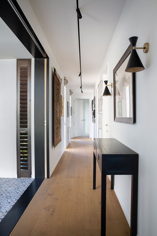 A long, bright residential hallway with pale timber flooring, white walls and ceiling, black-framed openings, and a series of black-and-brass wall sconces. A slim dark console table sits against the right wall beneath framed artwork, while patterned flooring is visible in an adjoining space at the left. Track lighting runs along the ceiling, creating a refined contemporary interior.