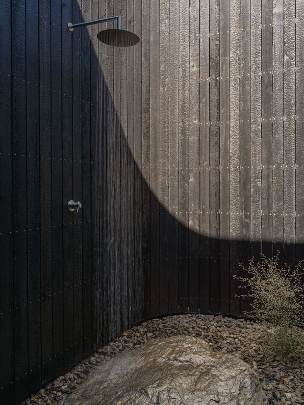 A dramatic outdoor shower mounted against a curved wall clad in dark, vertically ribbed timber boards. Water falls from a minimalist metal rain shower onto a natural stone and gravel ground surface, with strong sunlight creating a large curved shadow across the wall. Sparse planting appears at the edge of the frame.