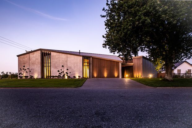 A low, single-storey contemporary building viewed from across a paved road at dusk. The facade combines warm timber cladding with pale rendered or masonry wall panels, tall narrow glazed openings with vertical timber screening, and a shallow pitched metal roof. Landscape lighting highlights the entry and facade, while a driveway leads to the recessed entrance. Mature trees and a neighbouring residential structure frame the property.