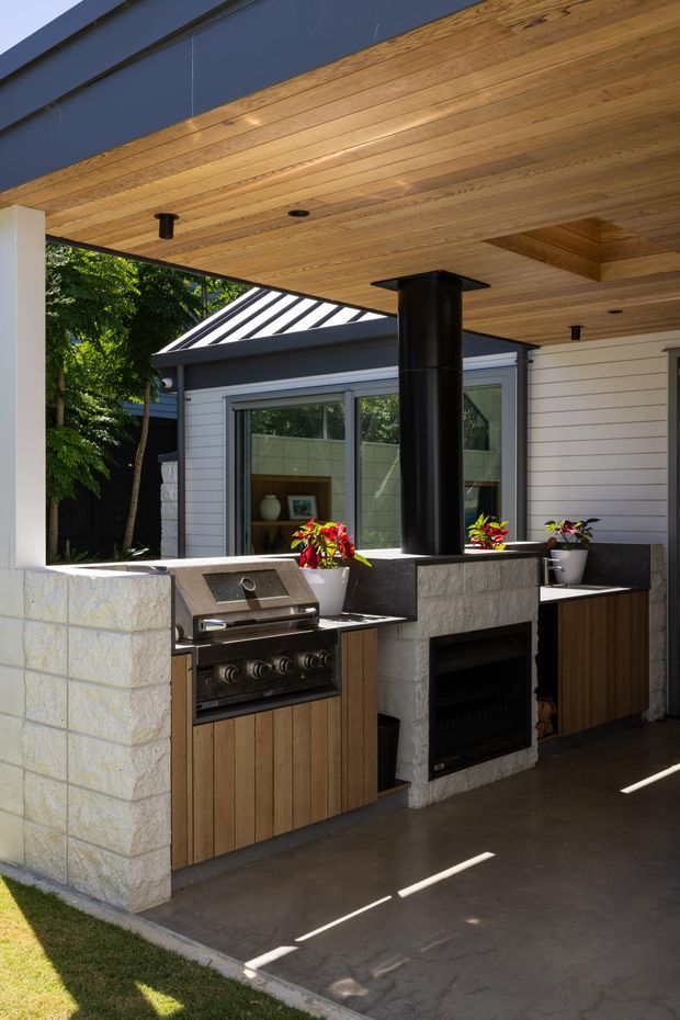 A covered outdoor entertaining area with a built-in outdoor kitchen, including a stainless-steel gas grill, dark stone or concrete benchtops, timber-fronted cabinetry, a central fireplace or pizza oven with a tall black flue, and potted plants. The space has a warm timber-lined ceiling with recessed downlights, pale masonry walls, dark cladding, and glazing overlooking greenery.