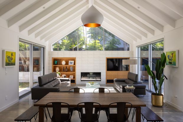 Bright open-plan living and dining space with a dramatic white vaulted ceiling and exposed painted rafters. Full-height glazing and glazed doors bring in abundant daylight and overlook greenery. The room includes a dark leather sectional sofa, matching lounge seating, a timber dining table with dark chairs, built-in timber shelving, a tiled feature wall with integrated fireplace and television, pendant lighting, and indoor planting.