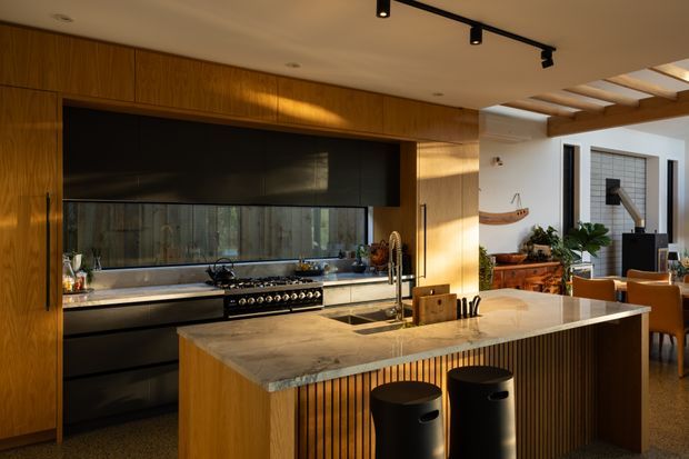 A sophisticated open-plan kitchen dominated by a large stone island with integrated sink, black stools, and vertically slatted timber detailing. The kitchen features extensive warm timber cabinetry, matte black overhead cabinetry and appliances, a reflective dark splashback, and a large range cooker. Track lighting and exposed timber ceiling beams extend toward an adjacent dining and living area, creating a warm, contemporary entertaining space.
