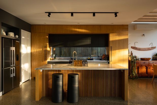A warm, contemporary kitchen with a central island featuring a pale stone benchtop, integrated sink and tap, and vertical timber slat cabinetry. Two black cylindrical bar stools sit at the island. The kitchen includes dark cabinetry, a large stainless-steel refrigerator, timber wall panelling, a dark rangehood, and a rear window or splashback opening. A dining area is visible through the opening to the right.