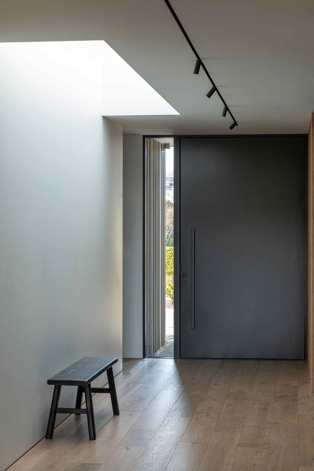 A minimalist residential entryway viewed from inside, with a large dark grey pivot door, narrow vertical sidelights, pale smooth walls, light timber flooring, a simple dark timber bench, and a black track light. A large geometric skylight or ceiling opening brings daylight into the space.