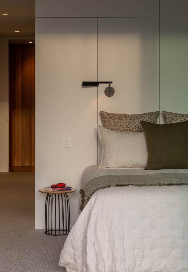 A softly lit, contemporary bedroom featuring a partially visible upholstered bed with layered neutral and olive bedding, a slim black wall-mounted reading light, and a small round timber-topped wire side table. The room has pale paneled walls, light carpet, and a warm timber-lined doorway leading to a hall or adjacent space.