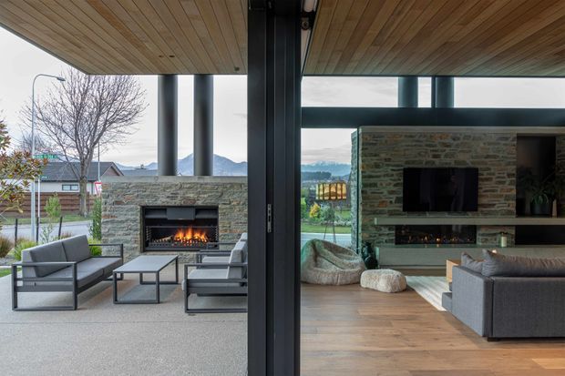 Contemporary indoor-outdoor living area centred on a substantial stacked-stone fireplace and matching built-in media wall. The image looks through wide dark-framed glazing or folding doors, with an open covered terrace on the left furnished with grey lounge chairs and a coffee table, and a warm timber-floored living room on the right with a sectional sofa, beanbag seating, and television cabinetry. Timber-lined ceilings continue across both zones, and the glazing frames a suburban mountain landscape.