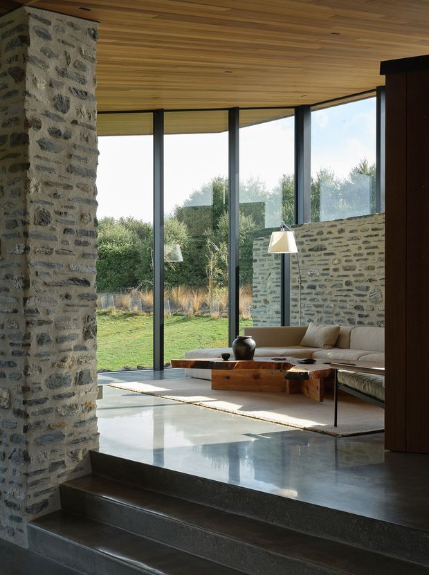 A refined, light-filled living room with full-height glazing overlooking a landscaped garden. Rough stone walls and a broad stone fireplace-like pier contrast with a warm timber-lined ceiling, dark slender window frames, polished grey flooring, and a low contemporary sectional sofa arranged around a substantial timber coffee table. Several steps create a raised living platform, while a dark timber built-in volume frames the right side.