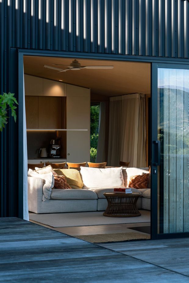 A warmly lit living room viewed from an exterior timber deck through a large open sliding glass door. The room features a pale sectional sofa, woven cushions, a dark woven coffee table, built-in timber cabinetry with a coffee station, full-height curtains, timber ceiling lining, and a ceiling fan. The exterior facade has dark vertical metal cladding, while the deck is finished in dark timber boards. A mountain landscape is visible through the glazing.