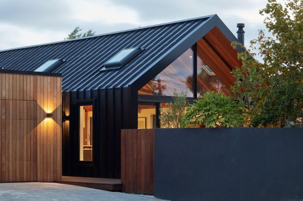 Exterior view of a compact contemporary house with a steep gabled roof clad in dark standing-seam metal, black vertical facade cladding, warm timber-lined gable soffits, and large triangular glazing. A timber gate and dark rendered boundary wall frame the entry, with concealed warm exterior lighting and planted screening.
