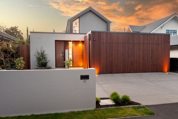 Contemporary residential street frontage at dusk, featuring a low white rendered boundary wall with black letterbox, a timber entry door glowing with warm interior light, a large vertical timber-slatted garage gate, concrete driveway, and a compact landscaped garden. The house behind has simple pitched roof forms and light-coloured cladding/render.