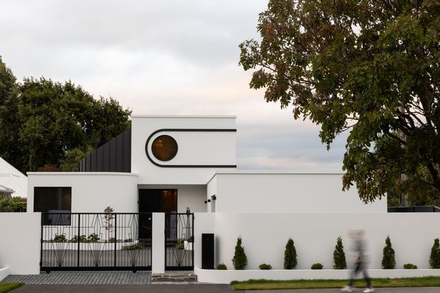 Street-facing view of a minimalist white residential house with strong geometric forms, a flat roof, black metal fencing and gate, a distinctive circular window, and a landscaped front boundary with clipped shrubs. A blurred pedestrian is visible on the footpath.