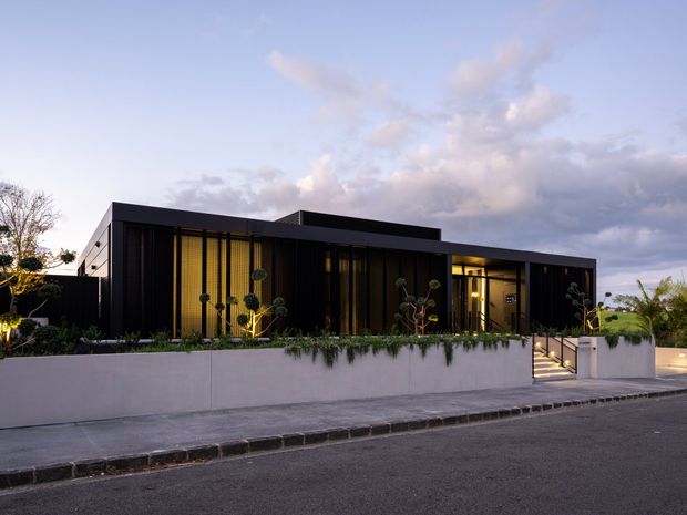 A contemporary single-storey residence viewed from the street at dusk. The home has a strong low-profile, flat-roofed form with dark cladding, extensive floor-to-ceiling glazing screened by vertical fins, and a recessed illuminated entry. A pale rendered boundary wall with integrated planting and a short stair leads from the footpath to the house.