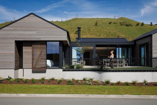 Single-storey contemporary home with dark horizontal timber cladding, a steep gabled roof over the left wing, and a low-pitched connecting wing opening onto a raised outdoor deck. Large glazed openings, timber screening, black-framed windows, and a black metal deck balustrade define the facade. The house sits against a grassy sloped site with simple landscaped planting along the street frontage.