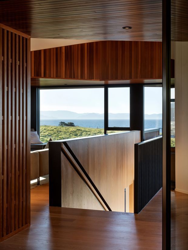 Interior view of a refined timber-lined stairway and landing overlooking a coastal landscape. The space features rich vertical timber battens, a warm timber slatted ceiling, dark-framed floor-to-ceiling glazing, timber flooring, and a sculptural stair with timber balustrade and black metal detailing. Ocean, grassy coastal terrain, and distant hills are visible beyond the windows.