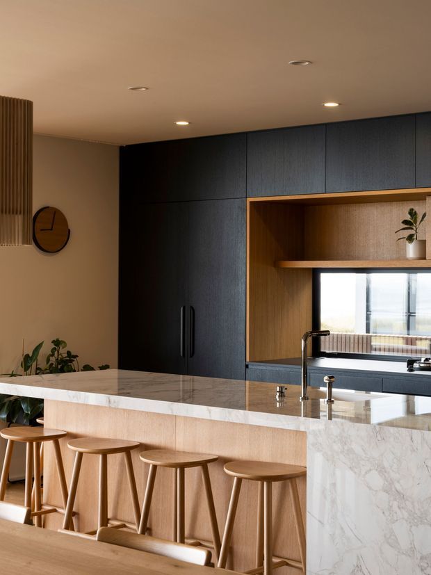 A warm, contemporary kitchen centred on a large marble-look island with an integrated sink and black tapware. The island has light timber-panelled cabinetry and seating for several people on simple timber stools. Behind it are tall dark blue-black wood-grain cabinets, a timber-lined open display niche, and a window above the rear benchtop. Neutral beige walls, recessed ceiling lights, indoor plants, and a round wall clock complete the refined interior.