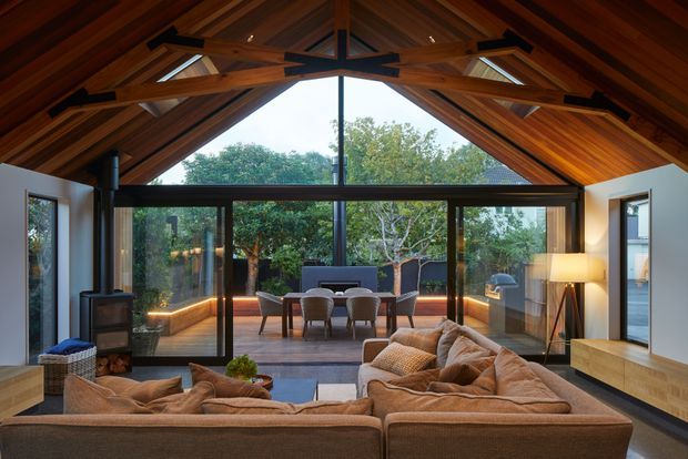 A warm, contemporary living room beneath a dramatic pitched timber ceiling with exposed structural beams and dark steel connections. The room opens through wide dark-framed glazing to a landscaped outdoor terrace, with a large sectional sofa, fireplace, media console, and layered ambient lighting. The timber ceiling and garden connection create a relaxed, indoor-outdoor character.