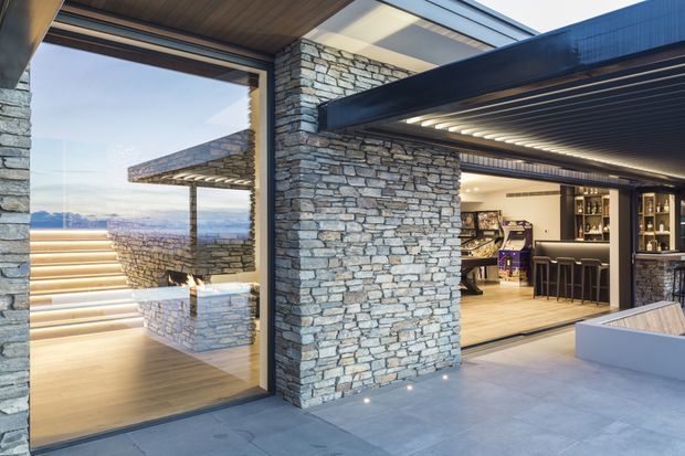 Contemporary outdoor entertaining area with extensive stacked-stone walls, timber soffits, dark metal detailing, and large glazed openings. The image looks from a covered terrace toward an integrated home bar and lounge, with illuminated steps and a distant landscape visible beyond the glazing. The space has a refined, resort-like character and strong indoor-outdoor connection.