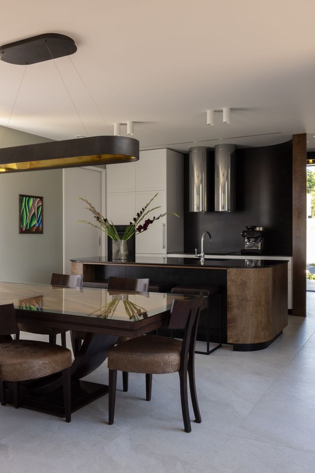 A refined open-plan kitchen and dining area with a dark timber dining table and upholstered dining chairs in the foreground. The kitchen features a large island with a dark benchtop, curved timber cabinetry, white full-height cabinetry, a dark feature wall, stainless-steel cylindrical rangehoods, and integrated appliances. Suspended sculptural pendant lighting and a glossy tiled floor contribute to the sophisticated contemporary interior.