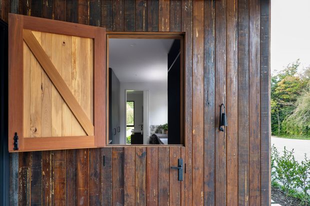 Exterior detail of a rustic timber-clad building with a large barn-style wooden shutter open beside a dark-framed doorway. The opening reveals a bright, modern interior beyond, while dense timber boards, black hardware, and natural weathered wood dominate the facade.