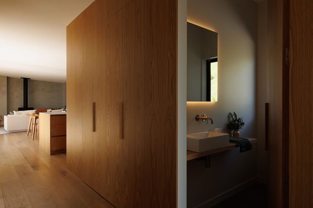 Warm contemporary interior showing a timber-lined built-in storage wall and concealed bathroom or powder room opening. The foreground is dominated by tall vertical-grain timber cabinetry with integrated handles. Through the opening, a compact bathroom is visible with a wall-mounted rectangular basin, brass tapware, mirror, dark benchtop or ledge, and a small window. Beyond the cabinetry is an open-plan living area with a pale sofa, kitchen cabinetry, stools, and timber flooring.