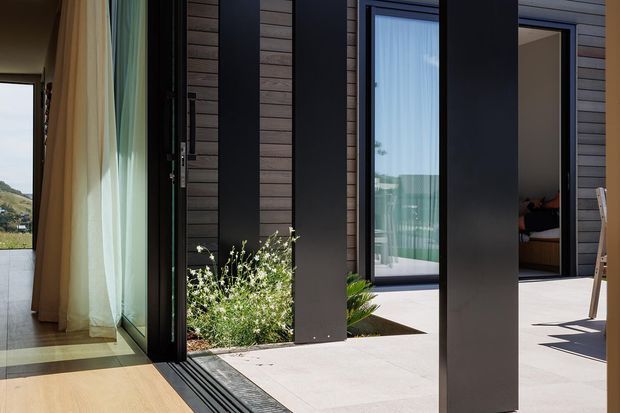 A contemporary indoor-outdoor threshold with large dark-framed glazed doors opening onto a pale paved terrace. Vertical dark cladding panels contrast with timber-toned interior flooring and sheer curtains, while a bedroom is visible through the open doorway. The composition emphasizes seamless indoor-outdoor connection, clean lines, and strong natural light.