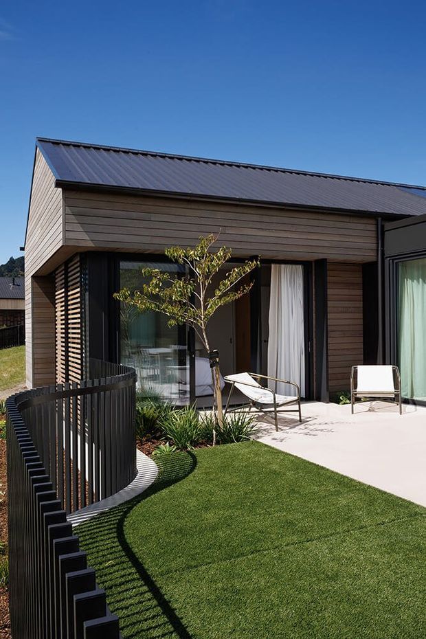 Exterior view of a contemporary single-storey building with a pitched corrugated metal roof, horizontal dark timber cladding, extensive black-framed glazing, and a pale paved terrace. A curved black slatted fence encloses a landscaped lawn with ornamental planting and outdoor lounge chairs.