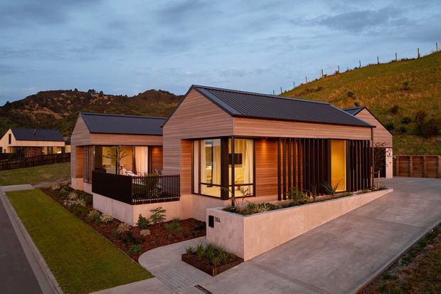 A modern single-storey residential house viewed from the street at dusk. The home has a low-pitched metal gable roof, warm horizontal timber-look cladding, extensive glazed openings, and a prominent vertical timber screen enclosing the front living area. Concrete paths and driveway surround landscaped garden beds, with rolling grassy hills visible behind the property.