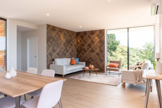 Bright contemporary open-plan living and dining area with pale timber flooring, a large natural timber dining table, soft upholstered seating, and a striking geometric dark timber feature wall behind the sofa. Floor-to-ceiling glazing opens to leafy outdoor views, while neutral furnishings and small orange accents create a warm, refined atmosphere.
