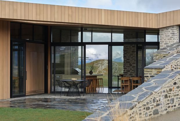 Contemporary mountain-facing residence with a broad glazed living and dining pavilion, dark-framed floor-to-ceiling windows, vertical timber cladding, and a rugged stone terrace and retaining wall. Indoor dining and seating areas connect directly to a stone-paved outdoor terrace, with mountain scenery visible through the glazing.