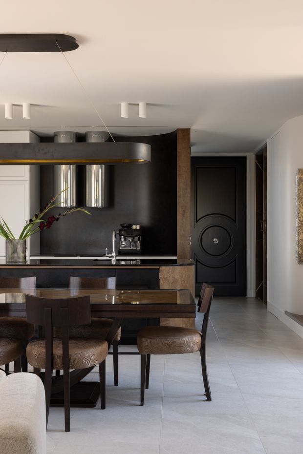 A refined open-plan dining and kitchen interior with a dark, sculptural kitchen island and integrated rangehood, dark cabinetry, metallic cylindrical splashback elements, and a rich wood dining table with upholstered dining chairs. A dramatic black paneled door and wood-clad structural column lead toward a hallway. Pale large-format flooring and light walls balance the darker joinery, while a linear pendant and recessed ceiling lights provide illumination.