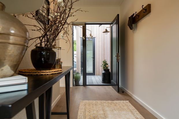 A warm, narrow residential entry hall with light painted walls, timber flooring, and a patterned runner rug. A dark console table with decorative ceramics and branches lines the left wall, while a timber coat rack is mounted on the right. Open glazed doors lead to a small exterior courtyard or deck with vertical timber cladding and potted plants.