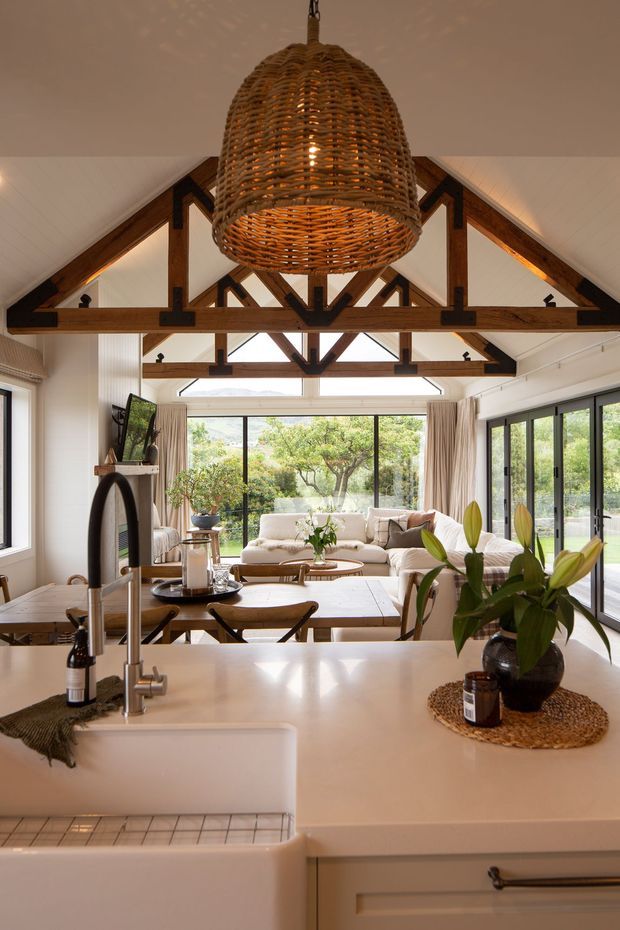 A warm, open-plan kitchen and living area viewed from behind a pale stone or solid-surface island. The kitchen features a deep white farmhouse sink, brushed metal tapware, light cabinetry and timber details. Beyond the island is a dining table and relaxed living room with large glazed doors and windows opening to greenery. The room has a vaulted white-lined ceiling with prominent exposed dark timber trusses and beams, along with a large woven pendant light.