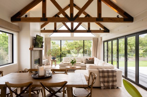 Bright open-plan living and dining room with large black-framed windows and glazed doors overlooking greenery. A long timber dining table occupies the foreground, while a cream sectional sofa, fireplace, coffee table and soft furnishings create a relaxed seating area. The defining architectural feature is the high pitched ceiling with exposed dark timber trusses and beams.