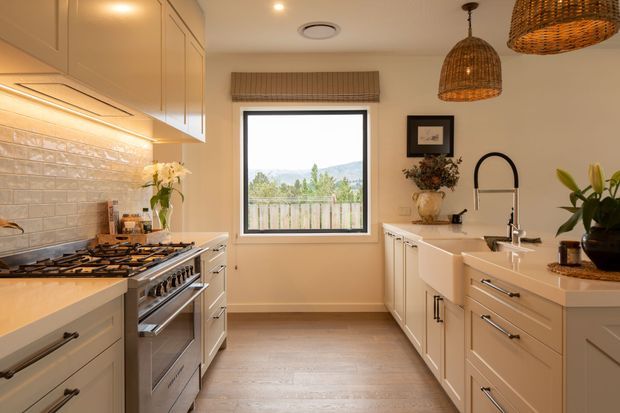 Warm, traditional-style kitchen with cream shaker cabinetry, a large central island, stainless-steel range, white farmhouse sink, and a glossy textured subway-tile splashback. Woven pendant lights, timber flooring, and indoor plants add natural warmth. A window provides a view toward a wooded, mountainous landscape.