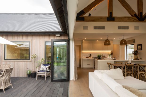 A bright open-plan living, dining and kitchen space opening directly onto a timber deck courtyard through large glazed doors. The interior features a white sofa, pale timber flooring, cream built-in kitchen cabinetry, a light island bench, timber dining furniture, woven pendant lights, and exposed stained timber roof trusses. The exterior deck has vertical timber cladding, a corrugated metal roof, outdoor seating and a large glazed opening.