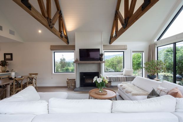 Bright open-plan living room with a large white sectional sofa, round timber coffee table, fireplace and television feature wall, and an adjacent dining area. Exposed timber trusses with dark metal connectors span the high ceiling, while large windows provide views of greenery and hills. The room has a relaxed, neutral, contemporary-rustic character.