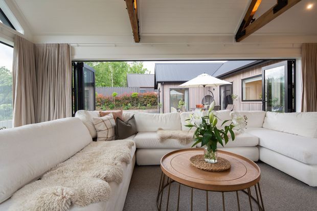 Bright contemporary living room with a large cream sectional sofa arranged around a round timber coffee table, soft textured throws, and a floral centerpiece. Full-height black-framed glazed doors open to a landscaped courtyard, while exposed dark timber ceiling beams contrast with the white panelled ceiling.
