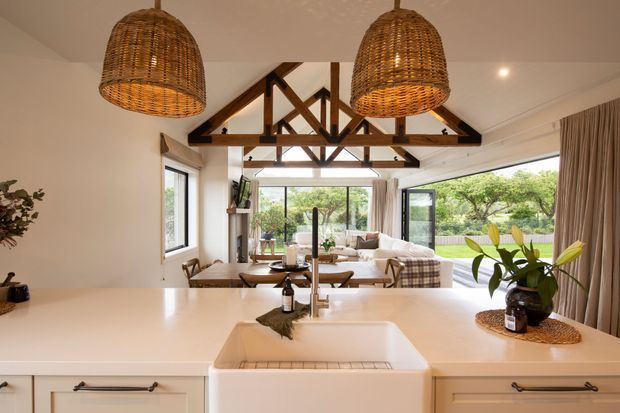 A bright open-plan kitchen looking through to a living area, with a large white island and farmhouse sink in the foreground. The room features exposed timber roof trusses, expansive glazing, woven pendant lights, white cabinetry, and warm neutral finishes. A garden is visible beyond the living space.
