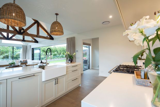 Bright, open-plan kitchen with white shaker-style cabinetry, broad white benchtops, a farmhouse sink, gas cooktop, timber-look flooring, and large windows overlooking greenery. Exposed dark timber roof trusses and woven pendant lights add a warm rustic character, while a connecting hallway and glazed door are visible beyond the kitchen.