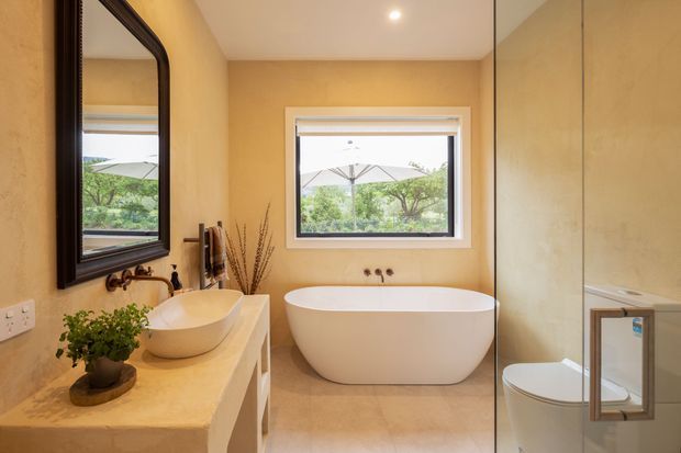Warm, refined bathroom featuring a freestanding oval bathtub beneath a framed window, a stone-look vanity with vessel basin, large dark-framed mirror, glass-enclosed shower, and toilet. The room uses beige plaster or stone-look finishes throughout with recessed ceiling lighting and views to greenery outside.