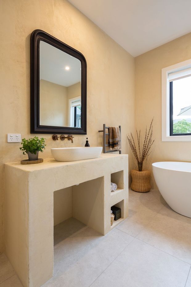 Warm, contemporary bathroom featuring a sculptural freestanding white bathtub, a textured beige plaster vanity with open shelving, an integrated white vessel basin, dark framed mirror, wall-mounted bronze tapware, towel ladder, and a light tiled floor. Natural light enters through a white-framed window, while woven and botanical accents add an organic touch.