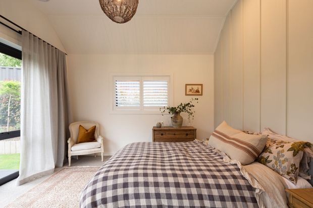 A warm, inviting bedroom with a neatly made bed, gingham check bedding, layered neutral and patterned pillows, and a vintage-style upholstered armchair. The room features pale paneled walls and ceiling, a large glazed opening with sheer curtains, plantation shutters, a timber dresser, woven pendant light, and soft neutral furnishings.