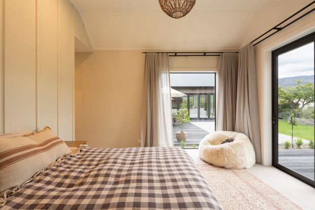 Bright, warmly styled bedroom with a large bed dressed in a brown-and-cream checkered coverlet, built-in full-height cabinetry along the left wall, soft beige curtains, and a sculptural pendant light. Floor-to-ceiling black-framed glazing opens to a deck and landscaped garden, with hills visible beyond. A plush cream lounge chair sits beside the window.