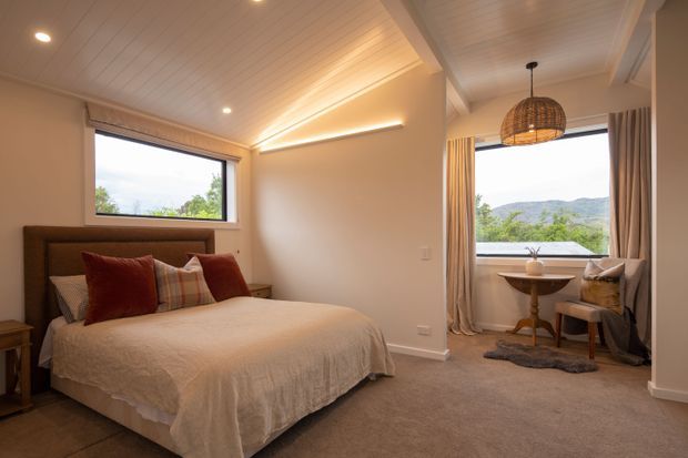 Warm, inviting bedroom with a large upholstered bed, layered neutral bedding and rust-coloured cushions. The room features carpeted flooring, cream walls, a white painted panelled ceiling with recessed lighting and a raised ceiling detail, plus a window-side sitting nook with a small round table and woven pendant light. Windows overlook a green, hilly landscape.