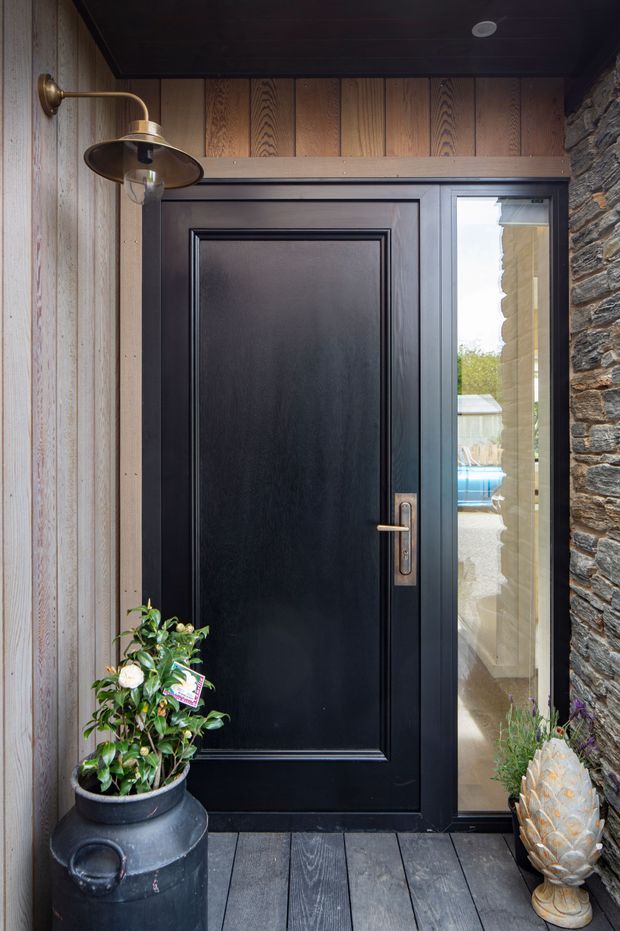 A refined residential entryway viewed from a covered exterior porch. The focal point is a tall black paneled front door with a narrow glazed sidelight and brass-toned hardware. Vertical weathered timber wall cladding, a dark timber ceiling, slate-like stacked stone masonry, and dark plank decking frame the entrance. A curved brass wall light, potted plants, and a decorative planter add character.