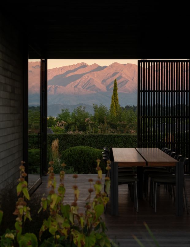 A dark, covered outdoor dining terrace frames a dramatic mountain landscape at sunset. A timber dining table with black chairs sits beside a vertical black slatted privacy screen, with timber decking, garden hedges, and planting extending toward the mountains. The composition has a moody, refined indoor-outdoor character with strong contrast between the shaded terrace and warm illuminated peaks.
