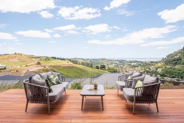 A sunlit outdoor terrace with a warm timber deck, two pairs of cushioned woven lounge chairs, and a small central coffee table. The terrace overlooks rolling green hills, a suburban coastal landscape, and the distant sea beneath a bright blue sky.