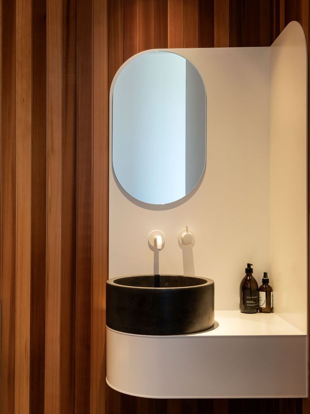 A compact, highly designed bathroom vanity vignette. A black cylindrical vessel basin sits on a white floating vanity with rounded corners, against a warm cream wall. Above it is a large rounded-rectangle mirror with a pale frame, flanked by two minimalist wall lights. The surrounding wall is clad in rich vertical timber battens, creating a warm, refined contrast with the sculptural white and black fixtures.
