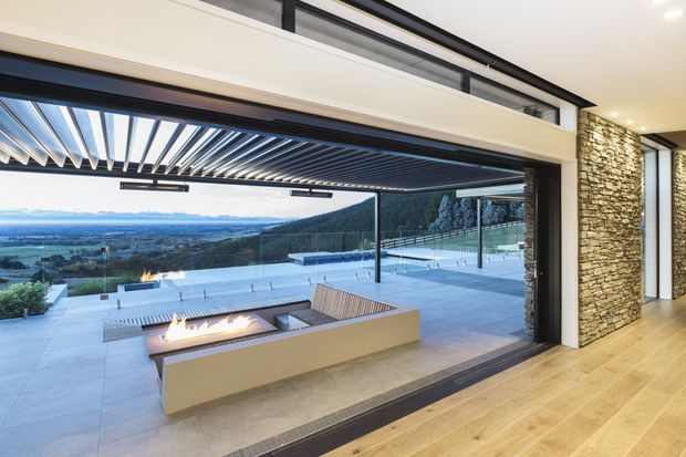 A contemporary indoor-outdoor living area opening onto a large terrace with a built-in linear fire feature and expansive rural landscape views. The opening is shaded by a dark louvred pergola, with frameless or slim dark glazing, pale timber flooring indoors, and a rough stacked-stone feature wall. The terrace has pale large-format paving, glass balustrades, and outdoor bench seating.